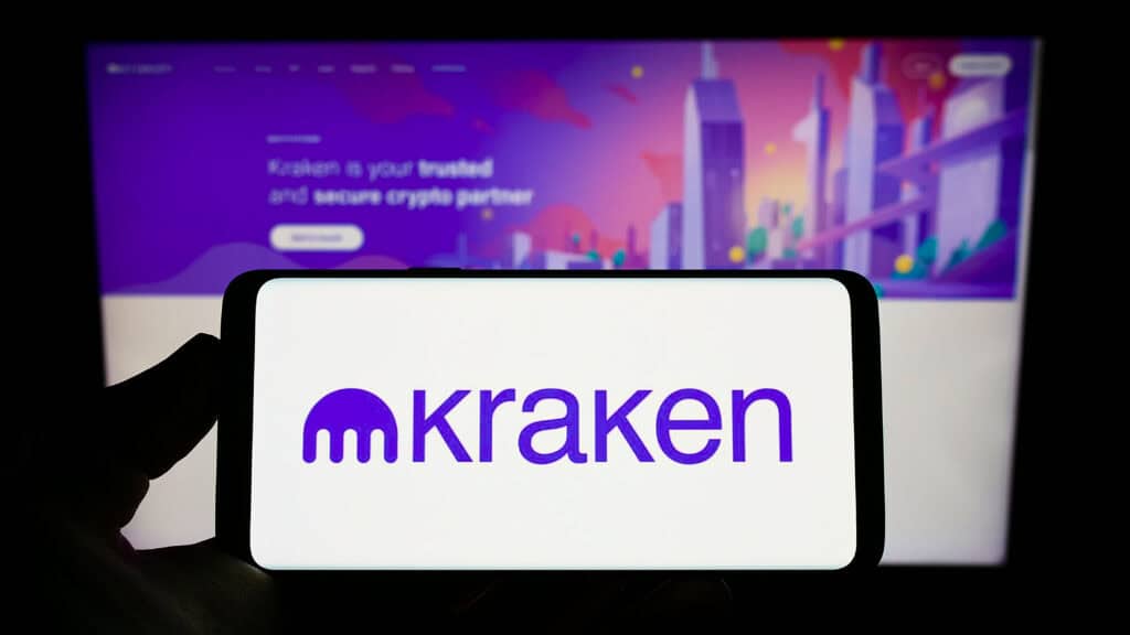 Person holding mobile phone with logo of Kraken Exchange on screen in front of web page.