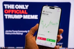 Man holding a phone displaying the official Trump meme logo in the Moonshot app, ready for purchase, with a blurred monitor in the background showing the meme's chart.