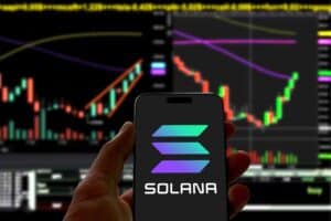 Smartphone with Solana logo is held in front of a screen that displays stock or cryptocurrency charts, bitcoin, investment, blockchain, trading, growth, succes