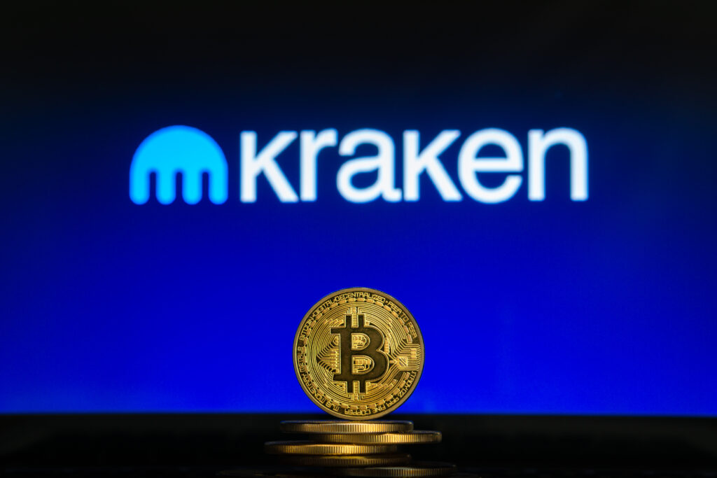 Bitcoin on a stack of coins with Kraken logo on a laptop screen.
