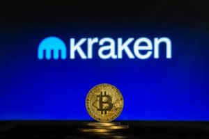 Bitcoin on a stack of coins with Kraken logo on a laptop screen.