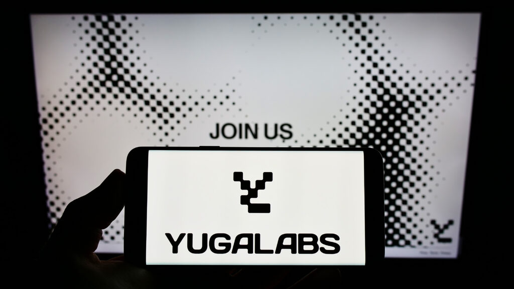 erson holding cellphone with logo of US NFT company Yuga Labs LLC on screen in front of business webpage.