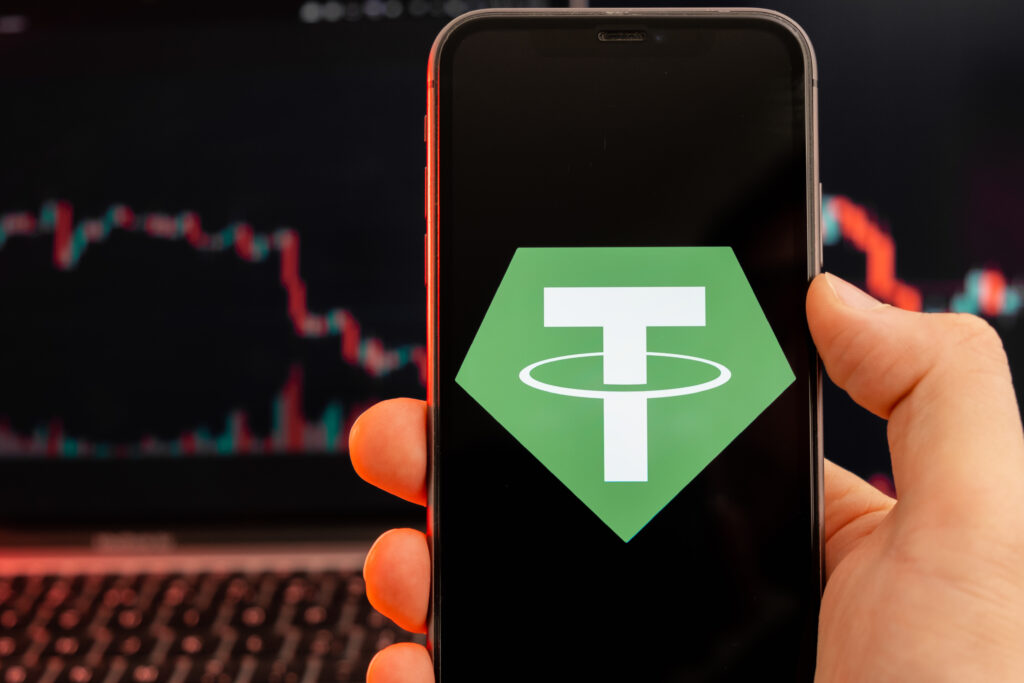 Tether USDT cryptocurrency logo on the screen of smartphone in mans hand with downtrend on the chart on a red light background