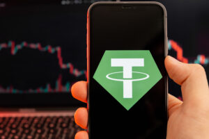Tether USDT cryptocurrency logo on the screen of smartphone in mans hand with downtrend on the chart on a red light background