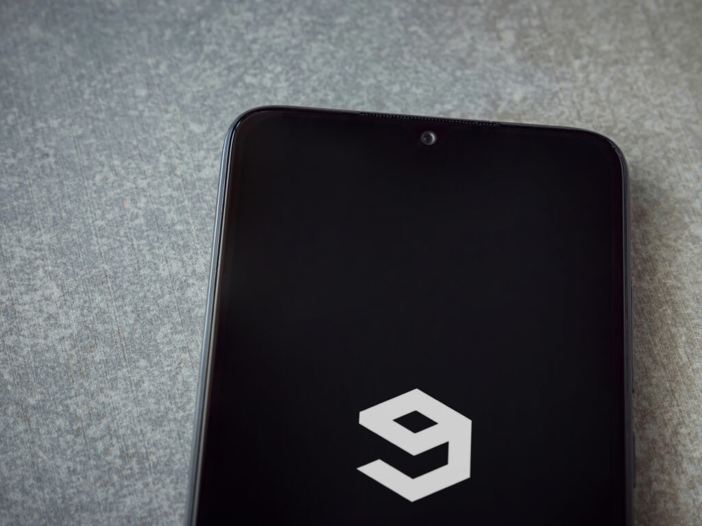 9GAG app launch screen with logo on the display of a black mobile smartphone on ceramic stone background.