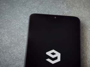 9GAG app launch screen with logo on the display of a black mobile smartphone on ceramic stone background.