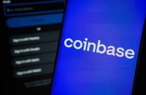 Coinbase logo is displayed on a smartphone screen, with the coinbase login page visible on a computer screen in the background.
