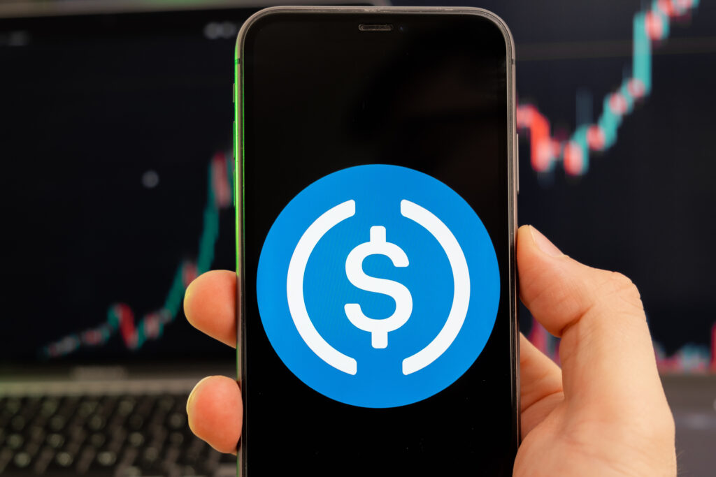 USD Coin cryptocurrency logo on the screen of a smartphone in mans hand with a growing trend on the chart on a green background.