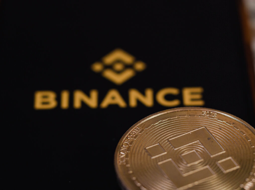 On screen mobile is the logo of the binance trading website. And there are bnb coins and dollars laying nearby.