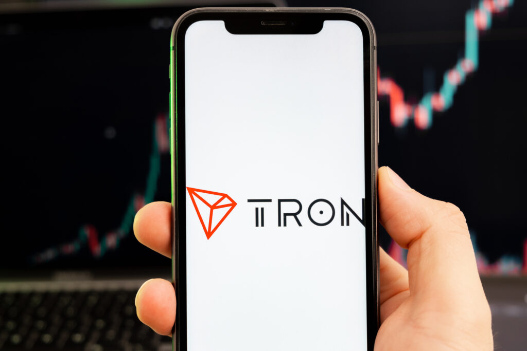 TRON TRX cryptocurrency logo on the screen of a smartphone in mans hand with a growing trend on the chart on a green background.
