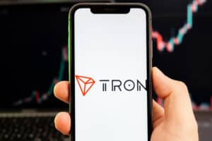 TRON TRX cryptocurrency logo on the screen of a smartphone in mans hand with a growing trend on the chart on a green background.