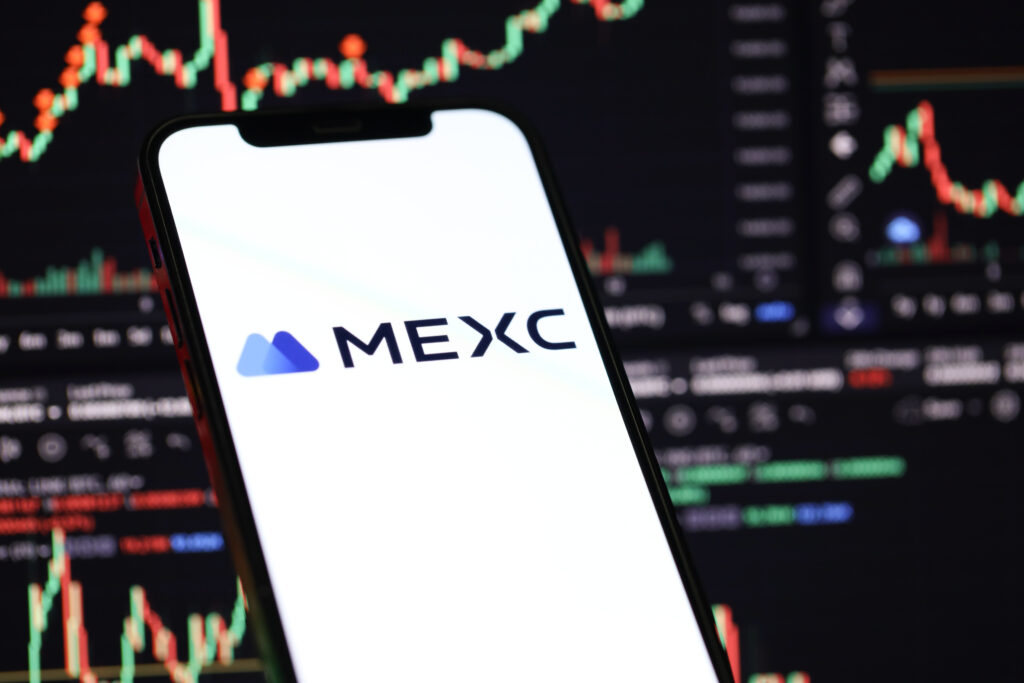 MEXC logo on iPhone display screen and crypto currency value charts.