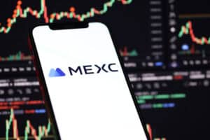 MEXC logo on iPhone display screen and crypto currency value charts.