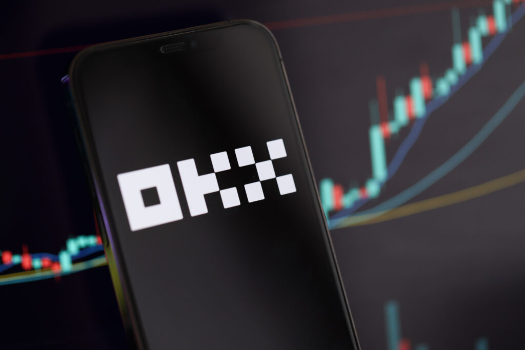 OKX logo on iPhone display screen and crypto currency value charts.