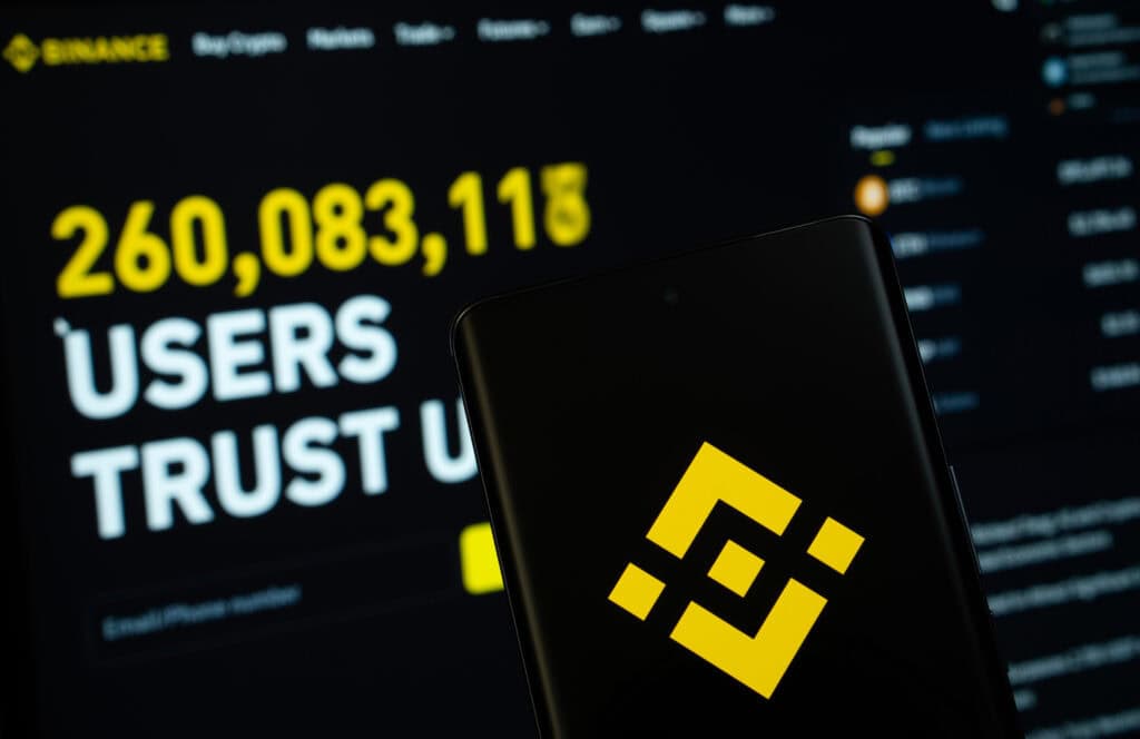 Binance logo is displayed on a smartphone screen, with the binance website visible on a computer screen in the background.