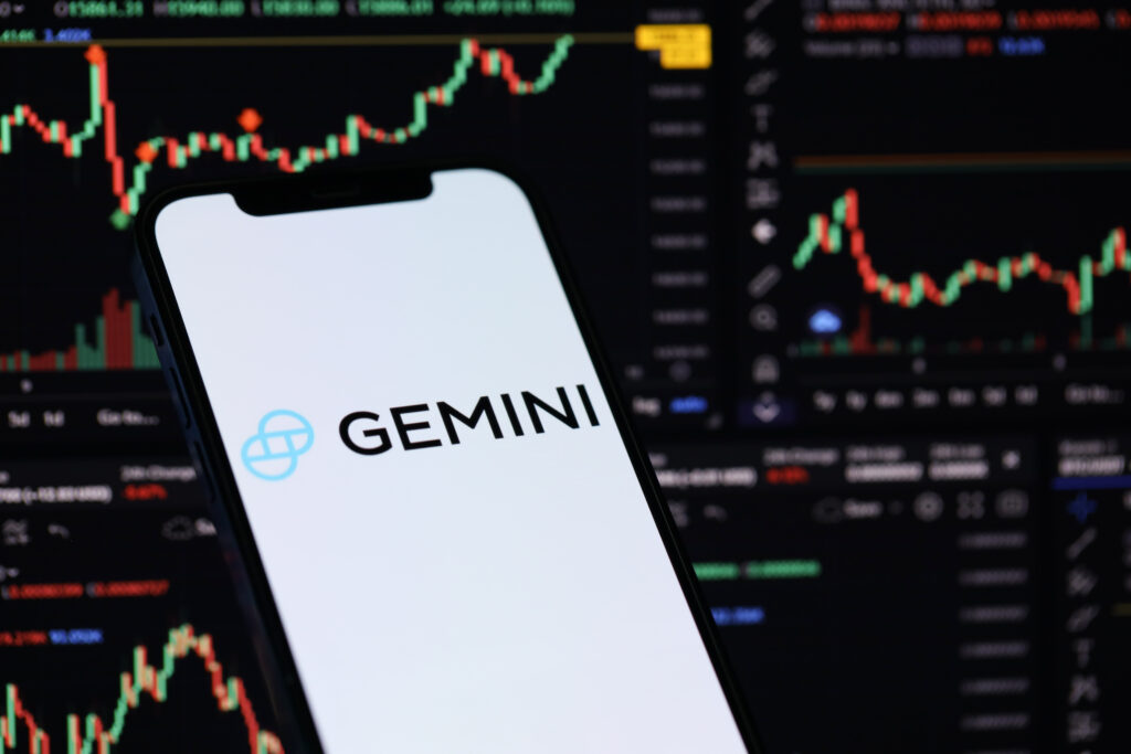 Gemini logo on iPhone display screen and crypto currency value charts