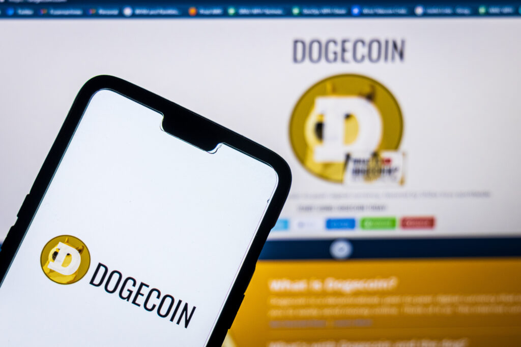 Dogecoin logo on a smartphone aganist dogecoin website on a pc in the background.