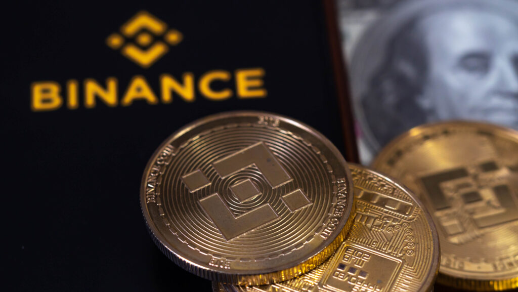 On screen mobile have logo of the binance trading website. And there are bnb coins and dollars laying nearby.