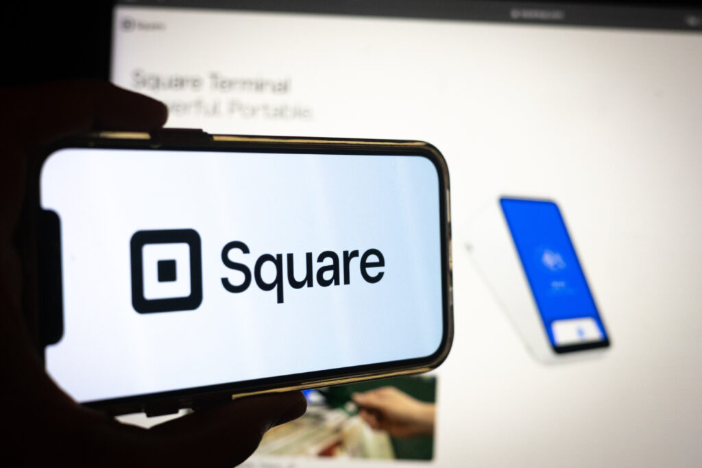 Silhouette of Hand Holding Phone with Square (SQ) Logo in Front of Square Payment Solutions Website on Computer Screen